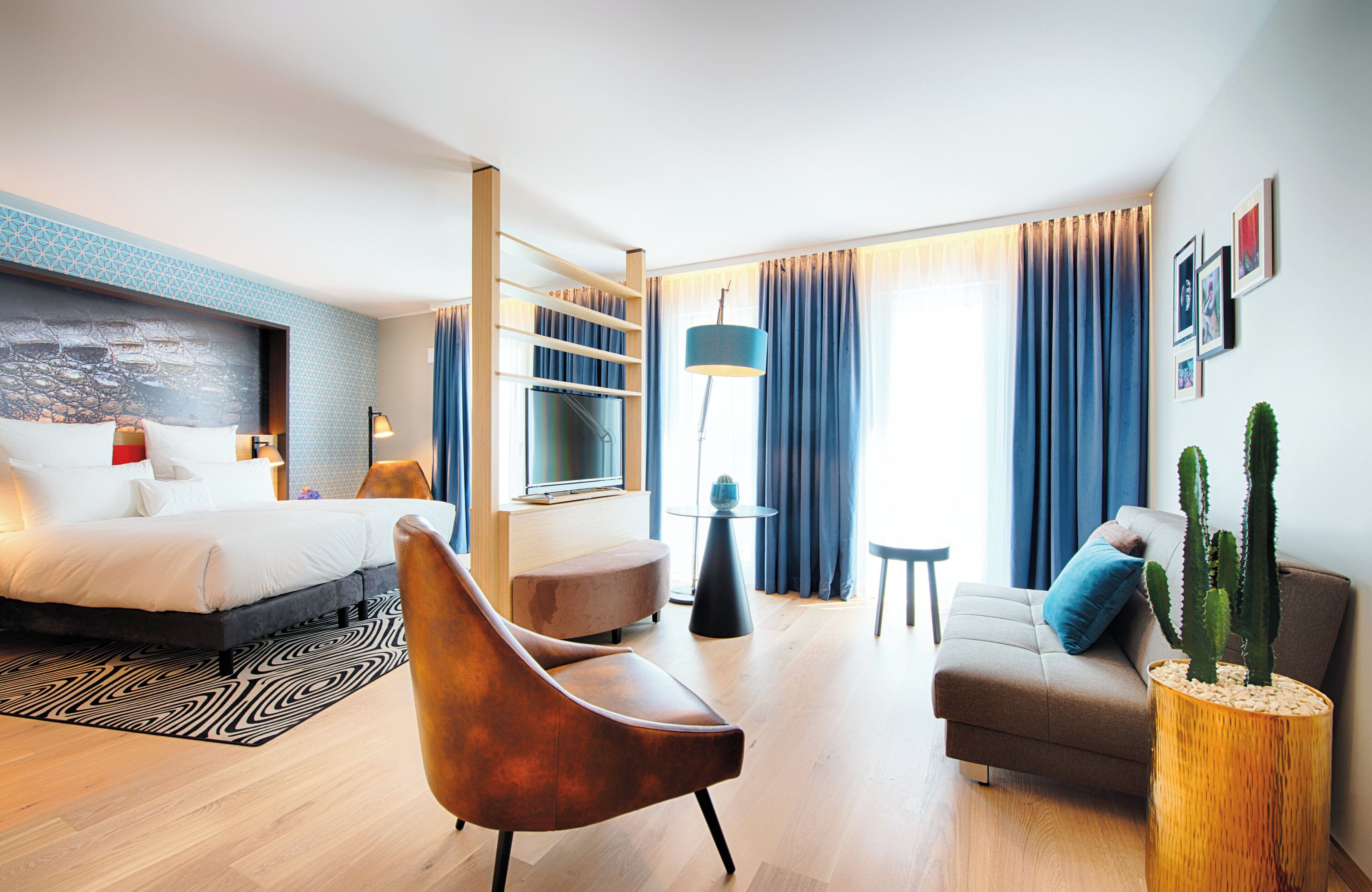 A modern hotel room with a bed, a sofa, a chair, a small table, a lamp, a cactus plant, and a large window with curtains.