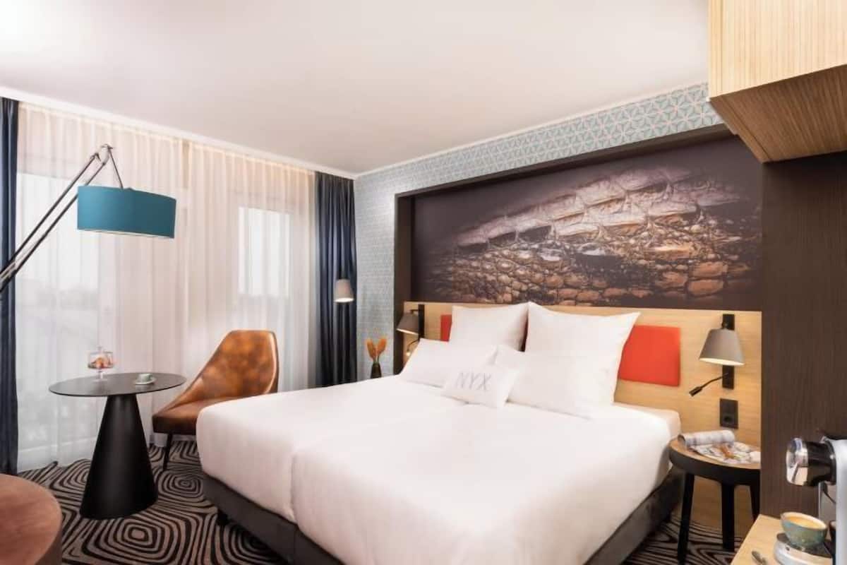 A hotel room with a bed, a desk, a chair, a lamp, and a painting on the wall.