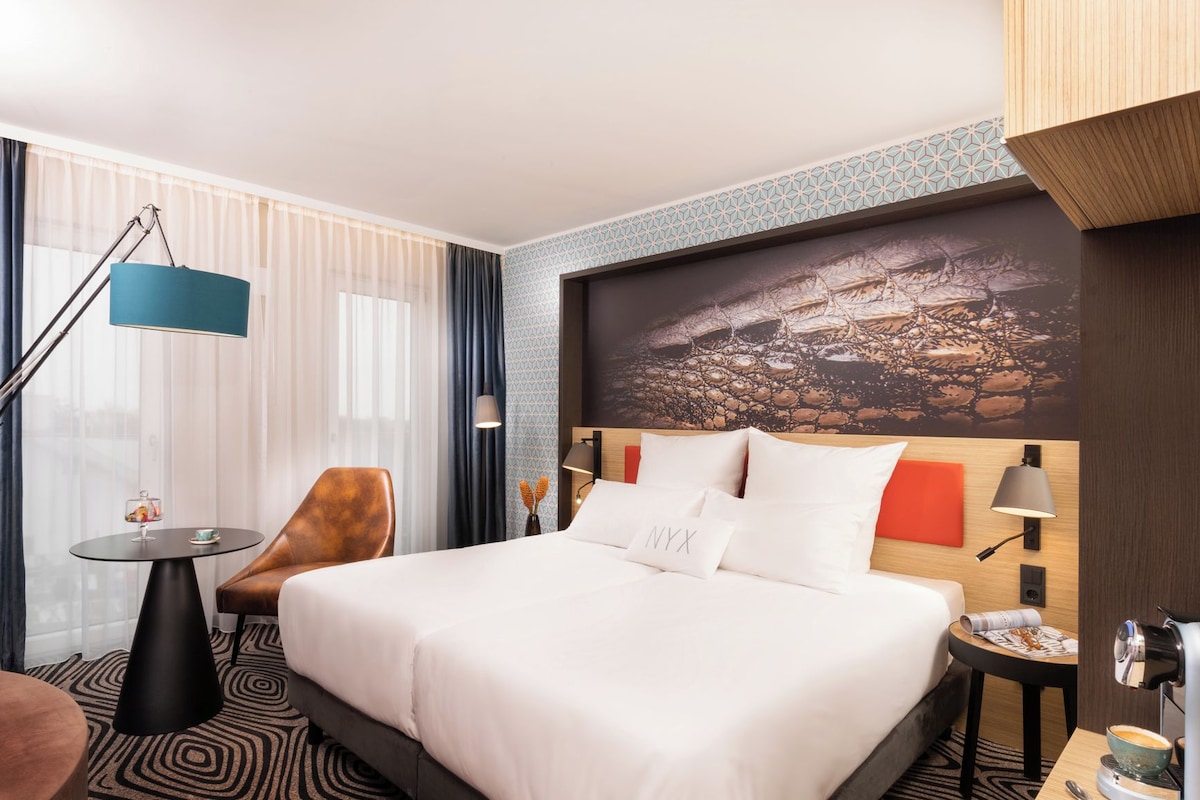 A hotel room with a large bed, a desk, a chair, a lamp, and a view of a cityscape.