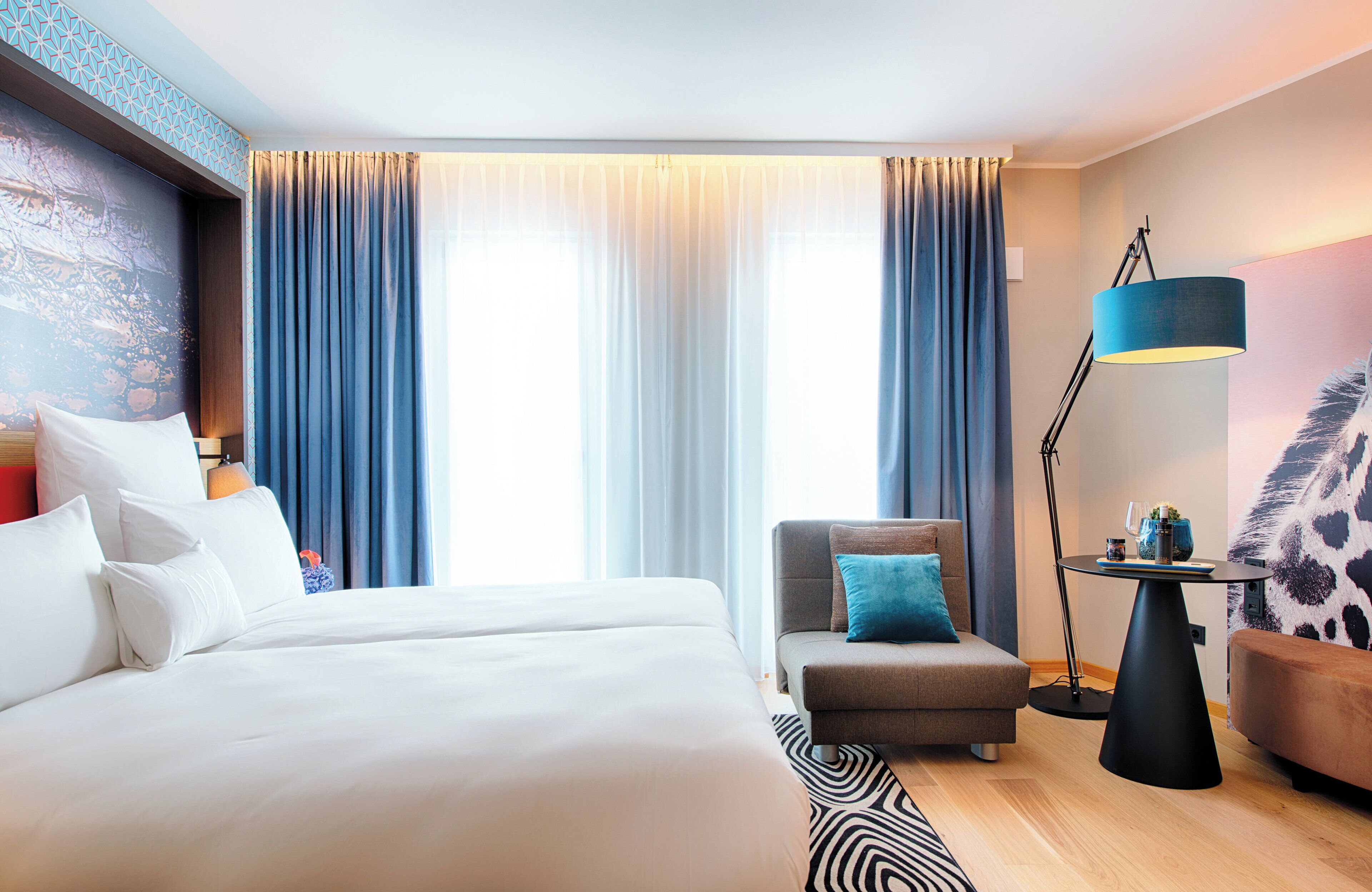 A modern hotel room with a large bed, a sofa, a small table with a lamp, and a view of the city through large windows.