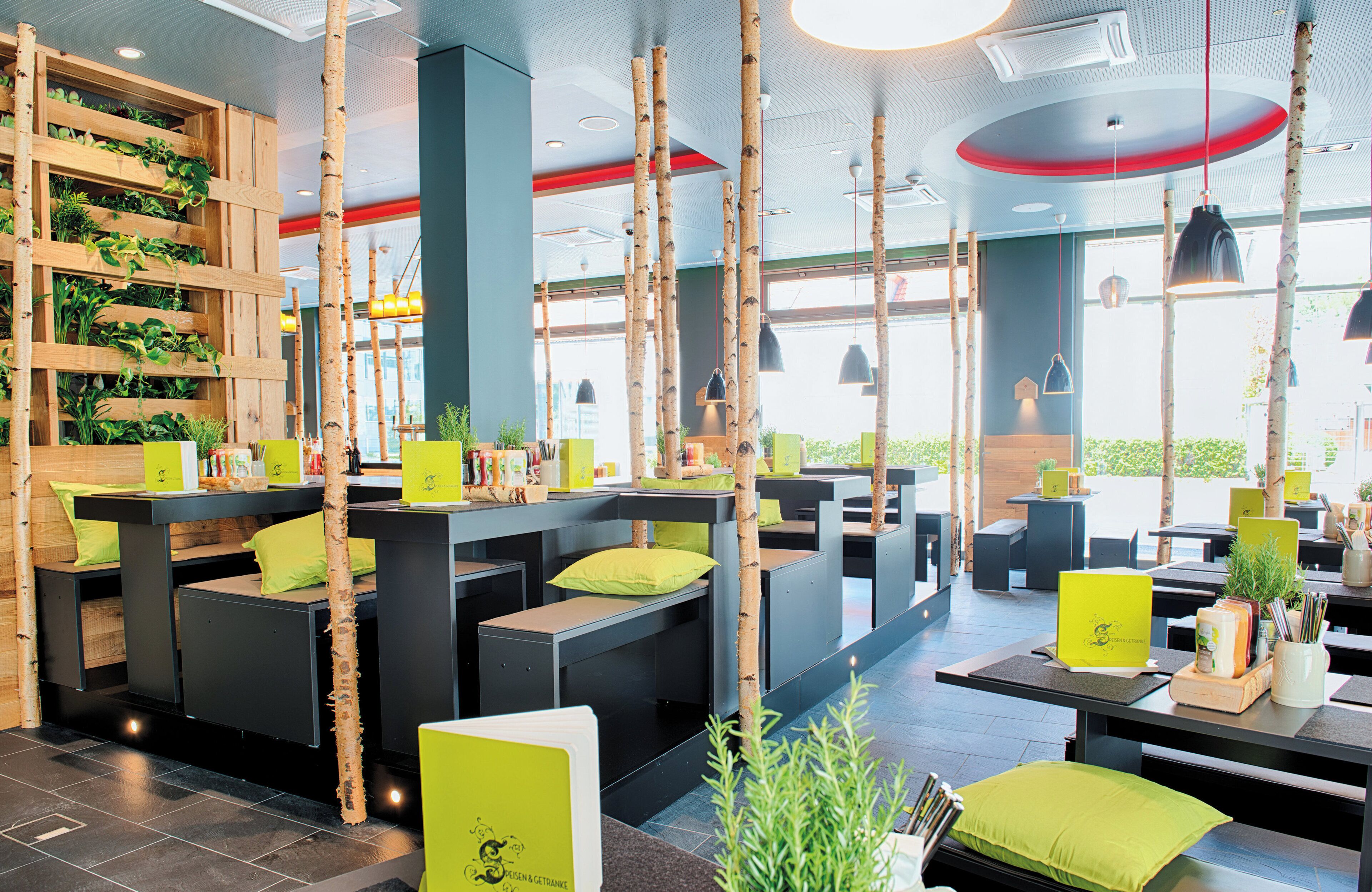 Un interior de restaurante moderno con altos paneles de madera, cojines verdes y luz natural.