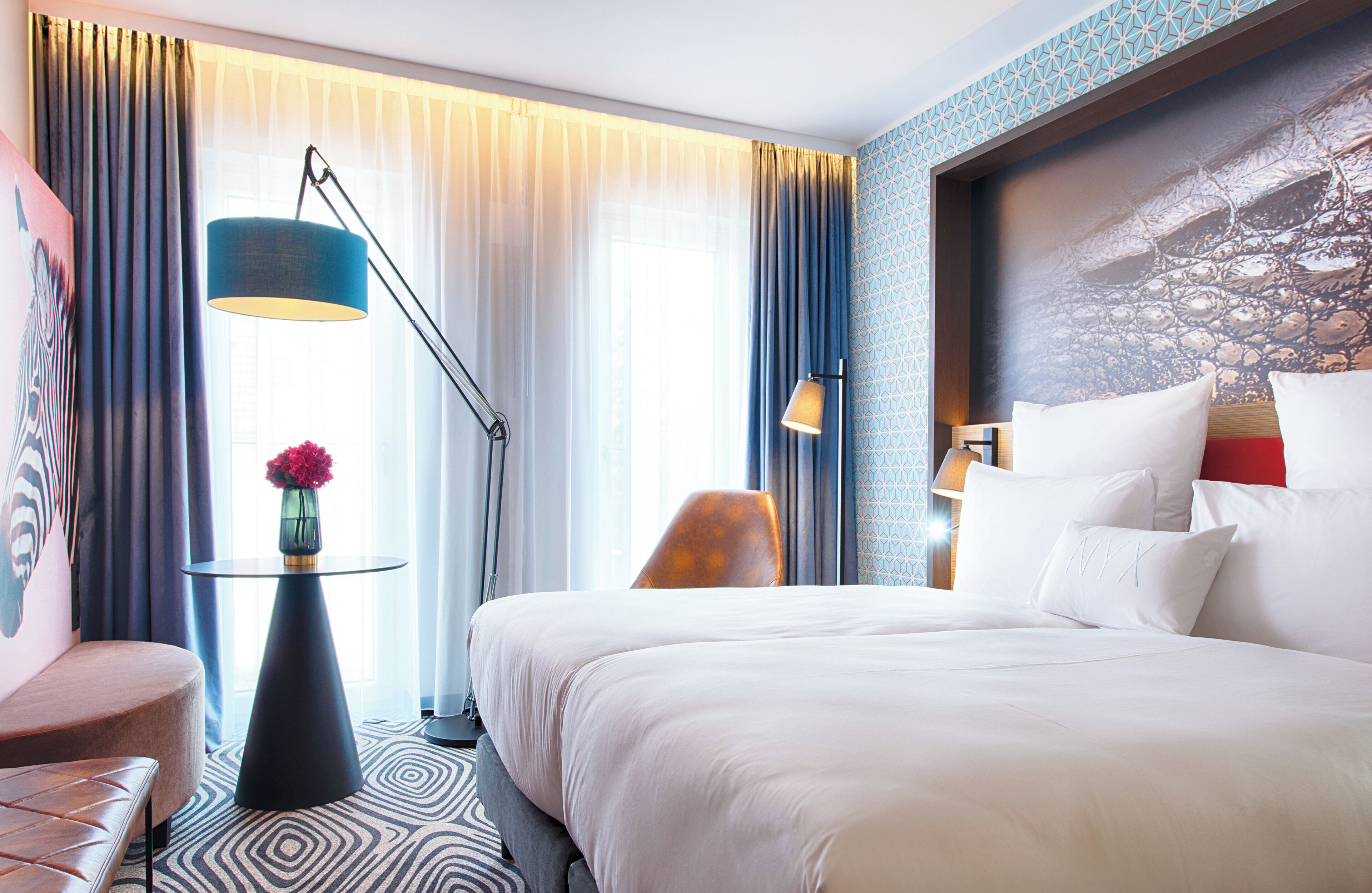 Ein Hotelzimmer mit einem großen Bett, einem runden Tisch mit Blumenvase, einer modernen Lampe und Blick ins Freie durch transparente Vorhänge.
