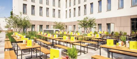 An outdoor seating area with tables and chairs, a building in the background, and greenery.