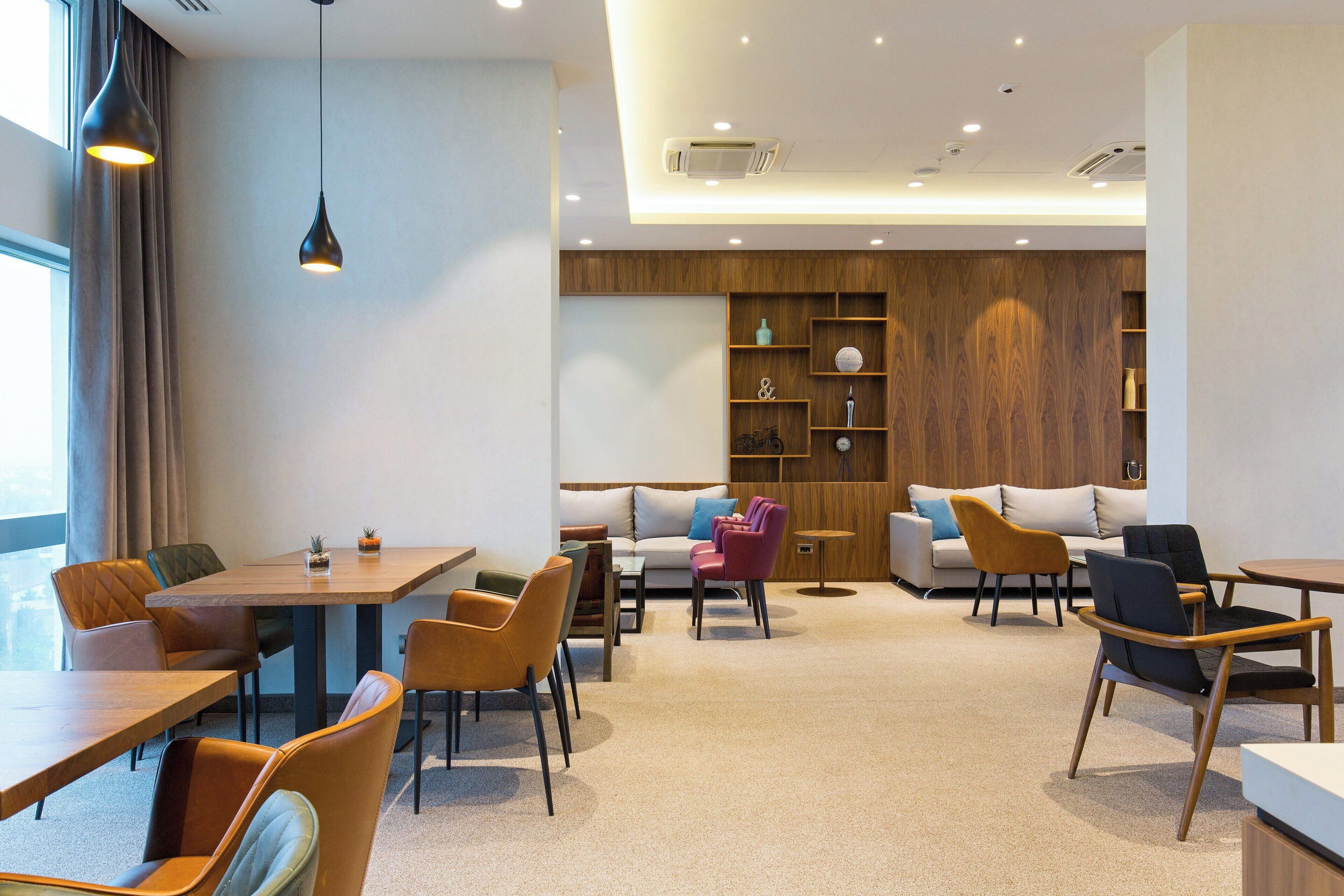 A modern hotel lobby with a wooden reception desk, seating area with sofas and chairs, and a large window with curtains.