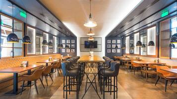 Un restaurante moderno con una mesa central rodeada de sillas, ventanas amplias y una pantalla de televisión en la pared.