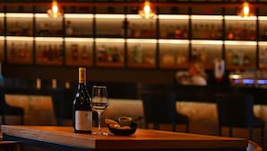 A bar with shelves of bottles, a bottle of wine, a glass, and a bowl of bread on a wooden table. - Sheraton Novi Sad (Novi Sad)