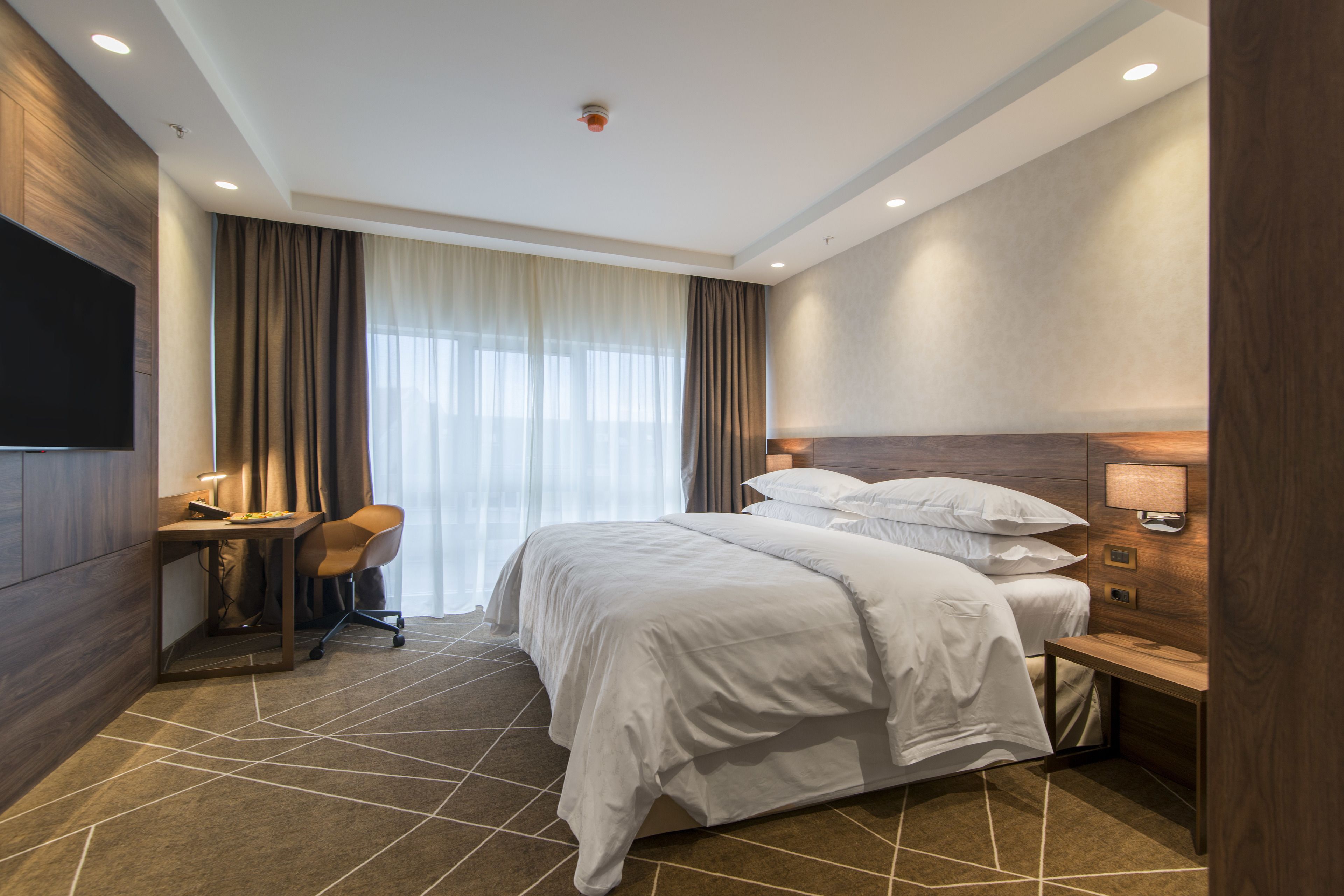 A hotel room with a large bed, a desk with a chair, a TV mounted on the wall, and a window with curtains.