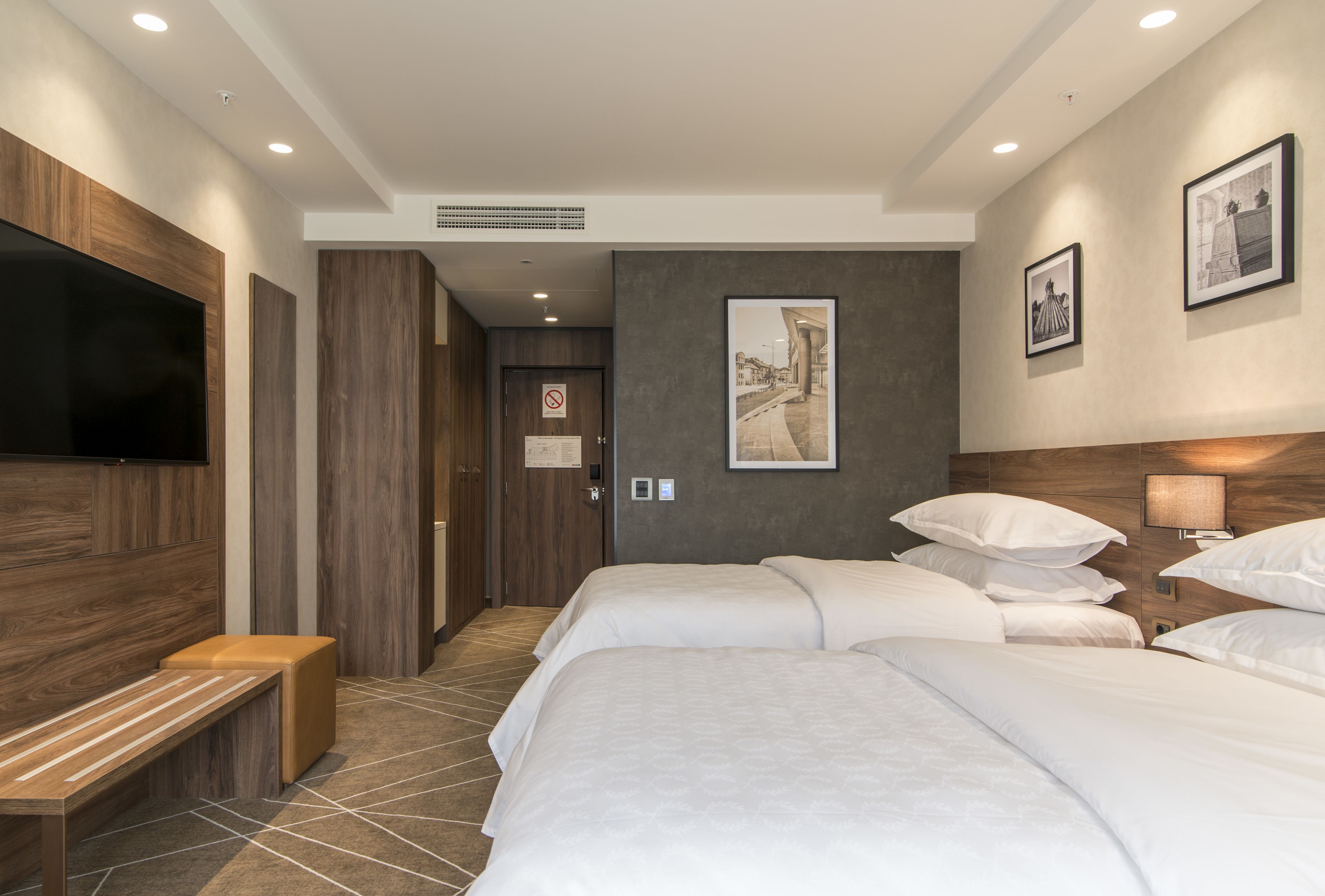 A hotel room with two beds, a TV, a bench, and artwork on the walls.