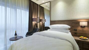 A hotel room with a large bed, a bedside table with a fruit bowl, a lamp, and a bottle of wine on a small table.