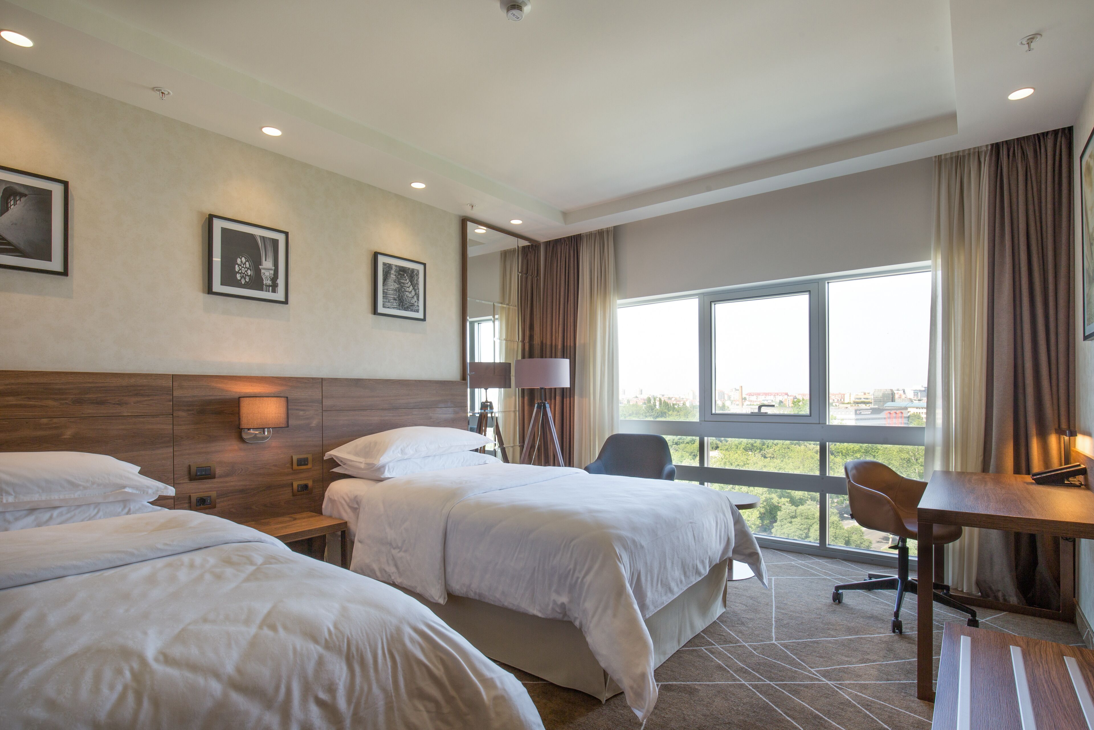 A hotel room with two beds, a desk, and a chair. There are framed pictures on the wall, a lamp, and a view of the city through large windows.
