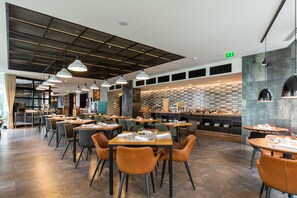 A modern restaurant with multiple dining tables and chairs, a kitchen area, and a counter with food displays. - Sheraton Novi Sad (Novi Sad)