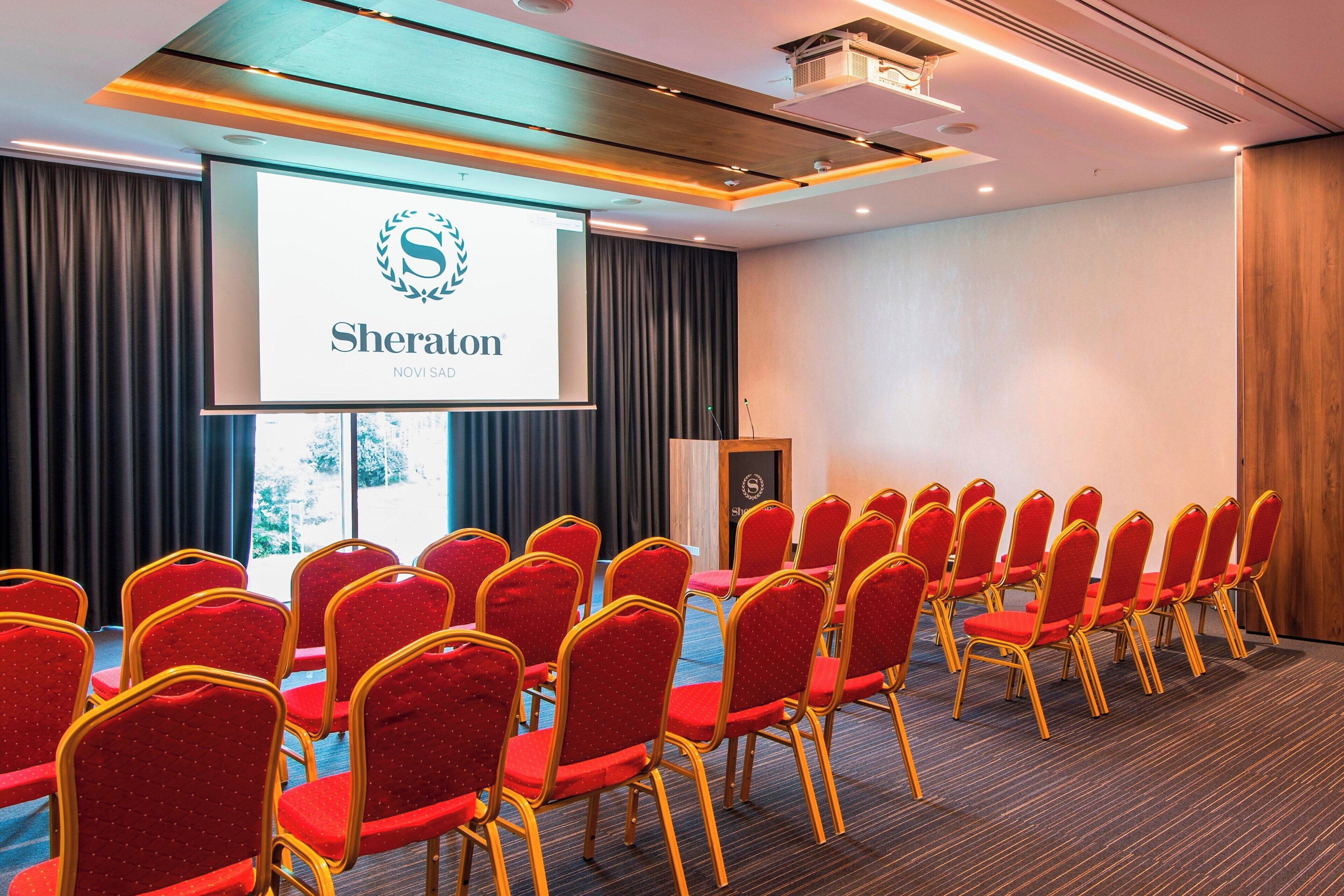 A conference room with red chairs, a podium, and a presentation screen displaying "Sheraton Novi Sad".
