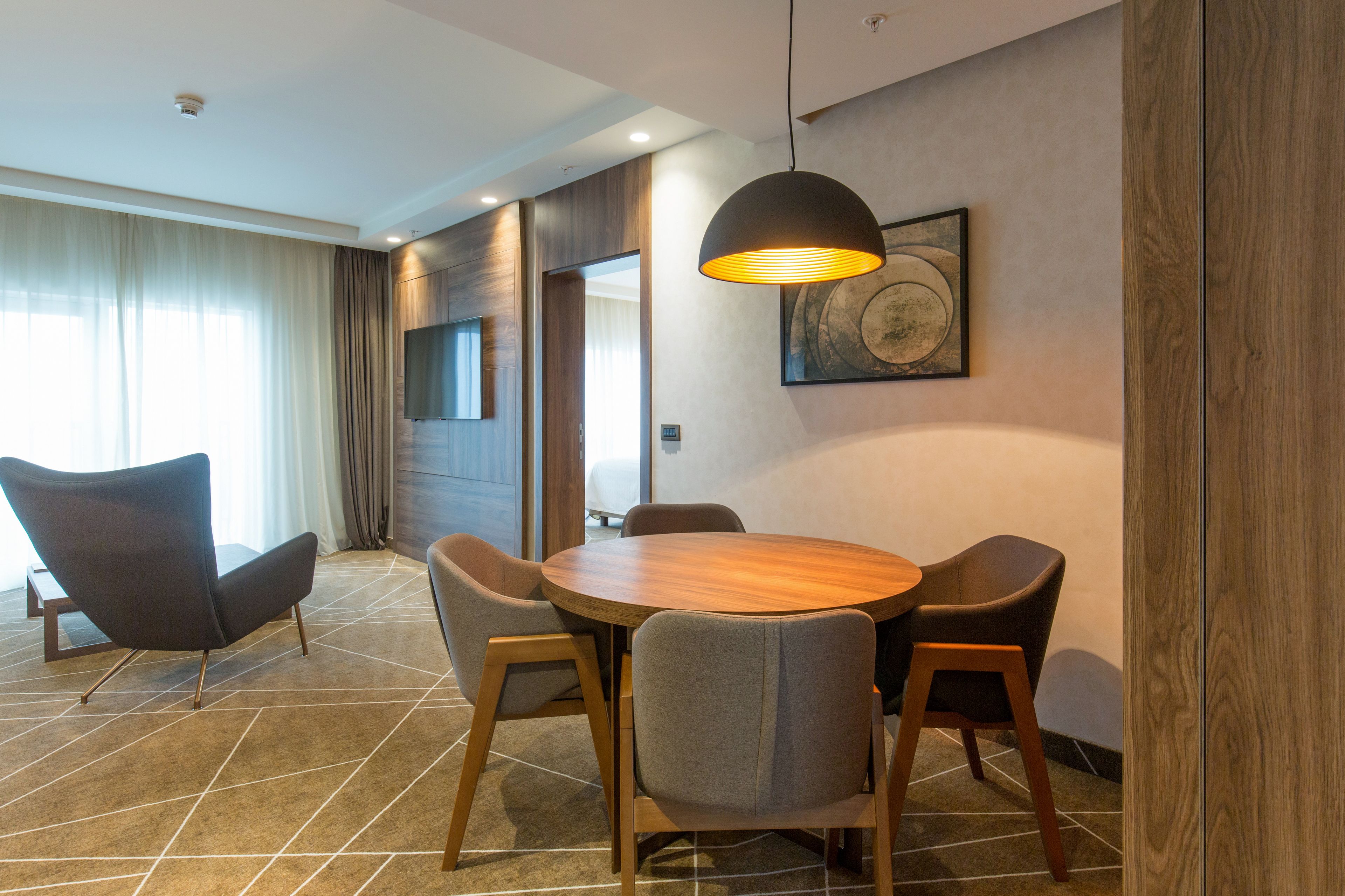 Una habitación de hotel moderna con una mesa de comedor redonda de madera, cuatro sillas grises, una lámpara colgante negra y un cuadro abstracto grande en la pared.