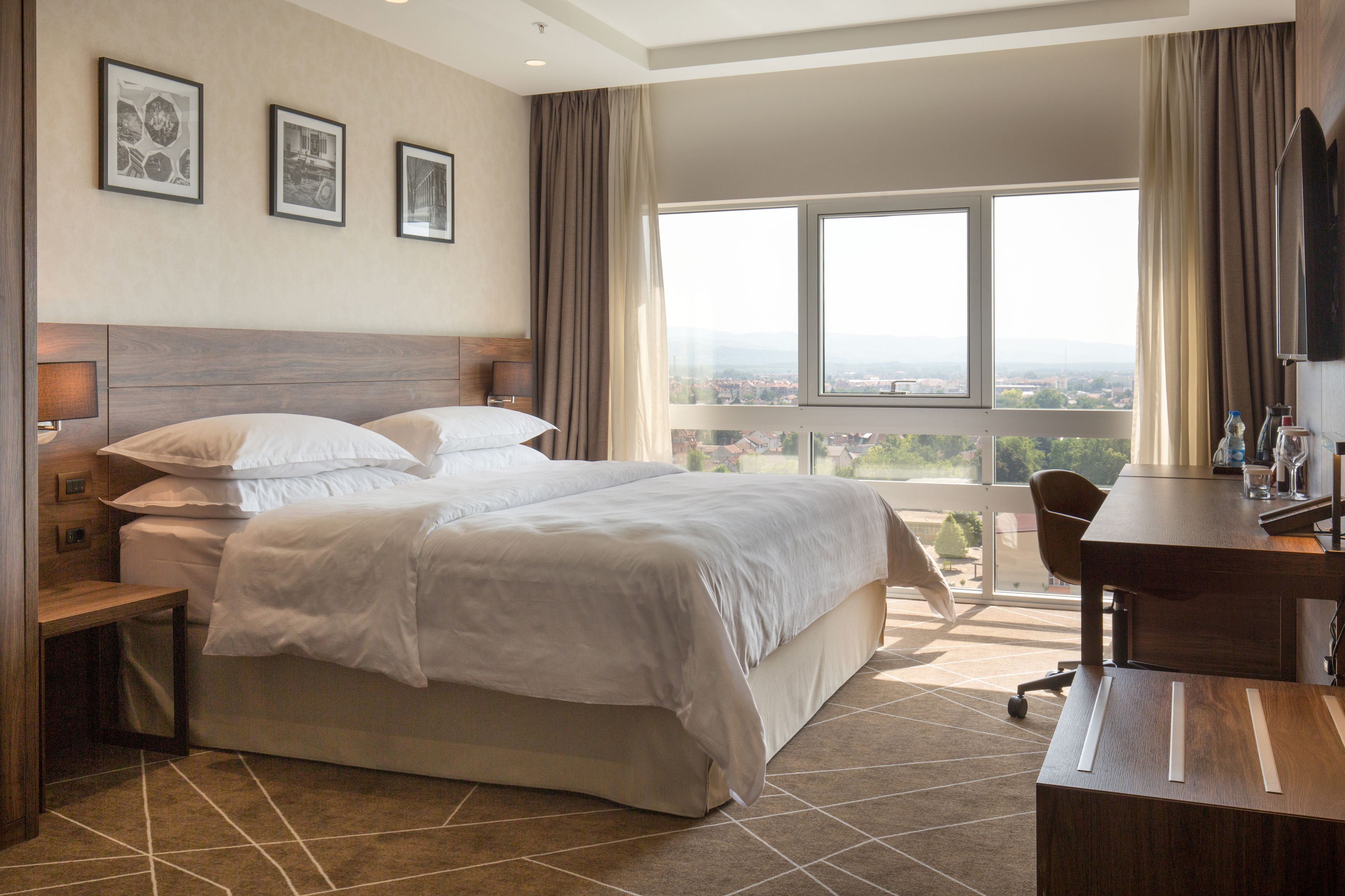 A hotel room with a large bed, a desk, and a view of the city through the window.