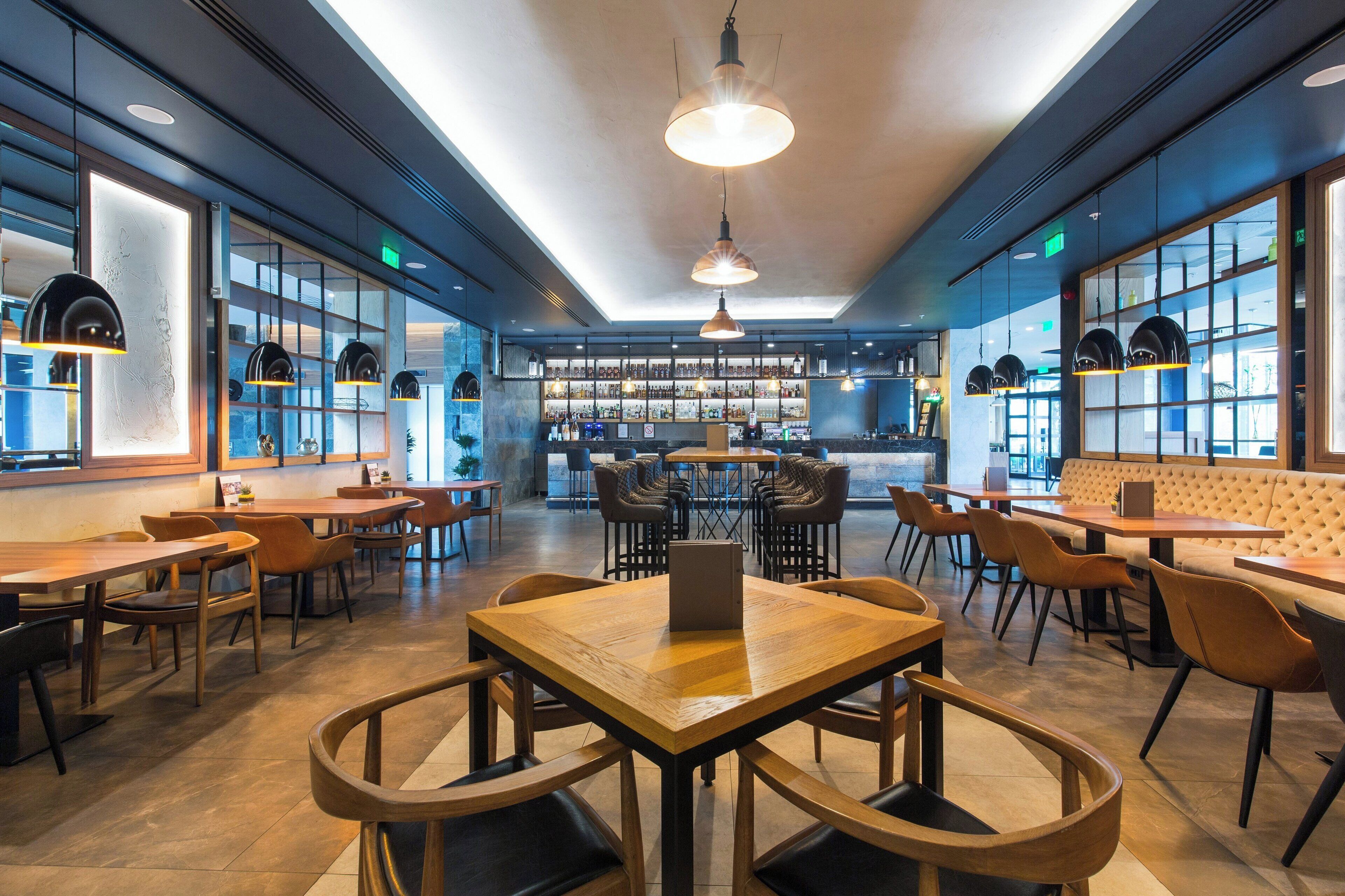 A modern restaurant with wooden tables and chairs, a bar area in the background, and large windows allowing natural light.