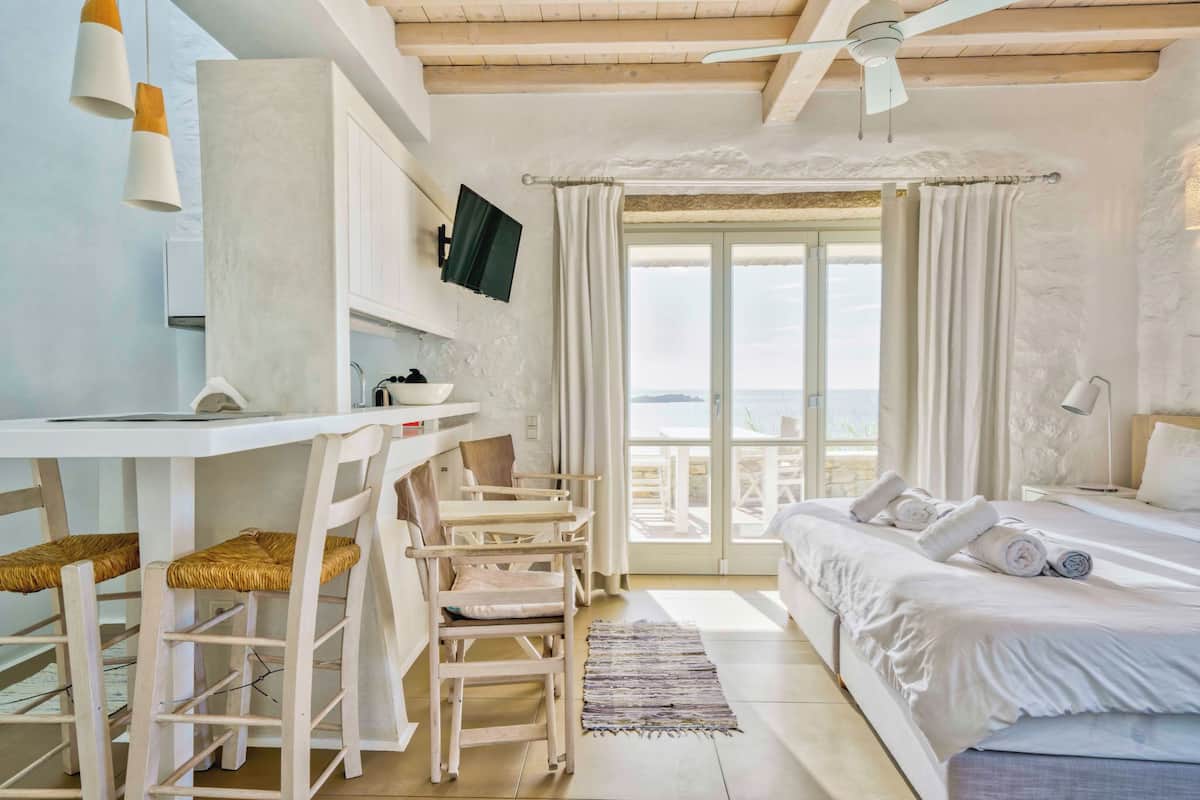 A bedroom with a bed, a desk, and chairs. There is a view of the outdoors through a large window.