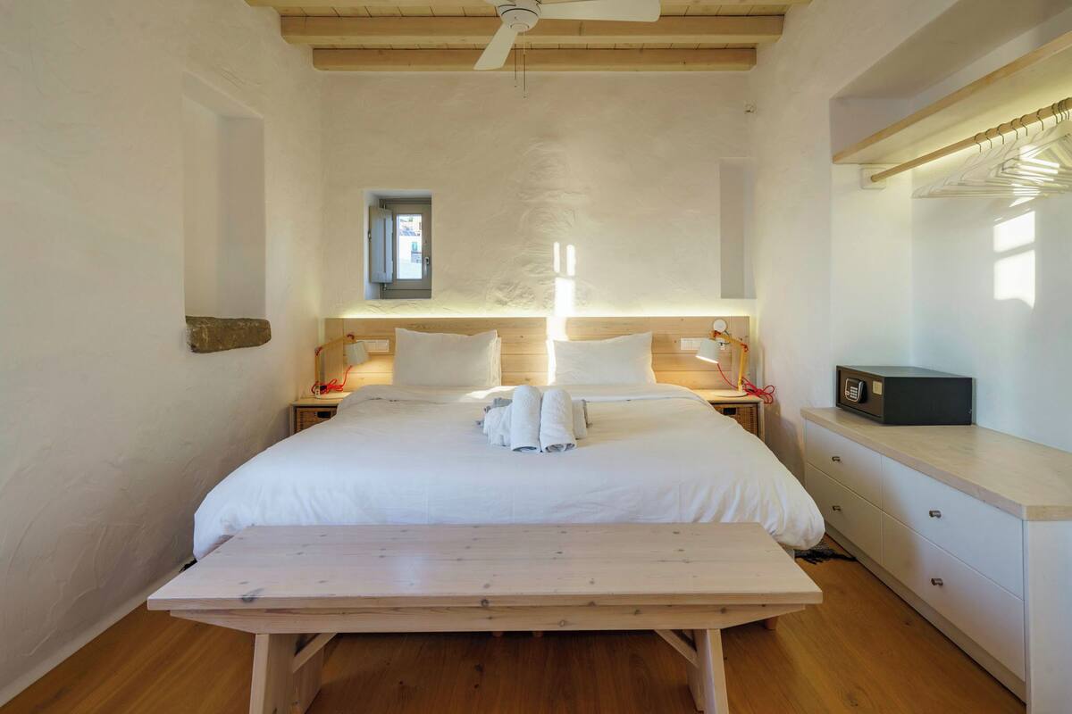 A bedroom with a wooden bed, white linens, a bedside table with a radio, and a wooden bench in front of the bed.