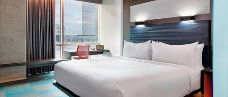 A hotel room with a large bed, a desk, a chair, and a view of the cityscape through the window.