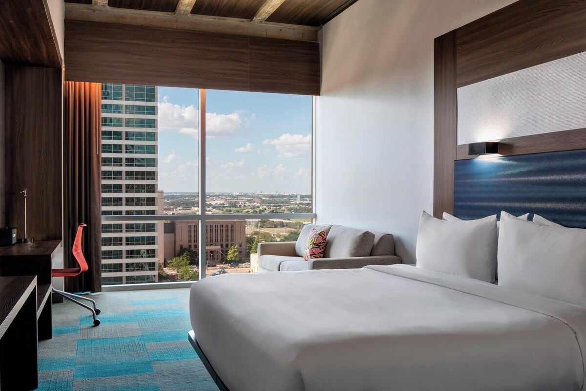 A hotel room with a large bed, a desk, a chair, and a view of a cityscape through a large window.