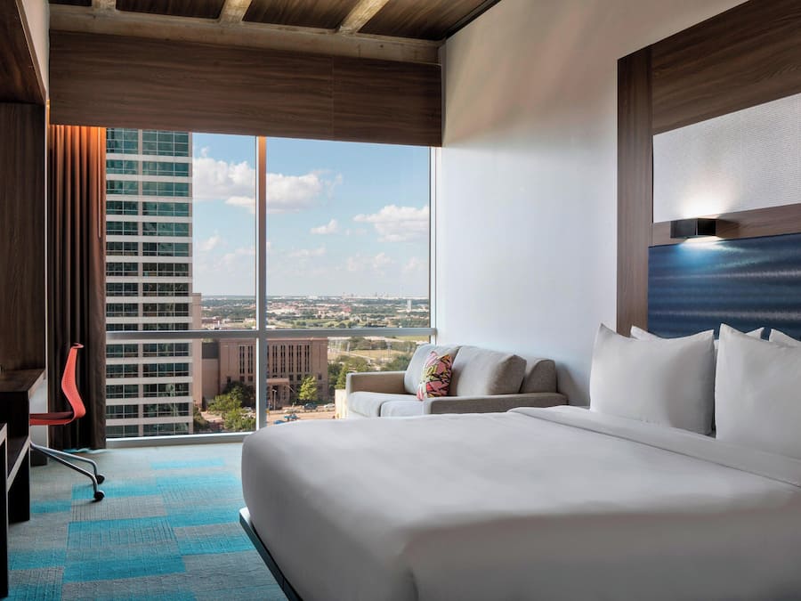 Une chambre d’hôtel avec un grand lit, un bureau, une chaise et une vue sur le paysage urbain, visible à travers une grande fenêtre.