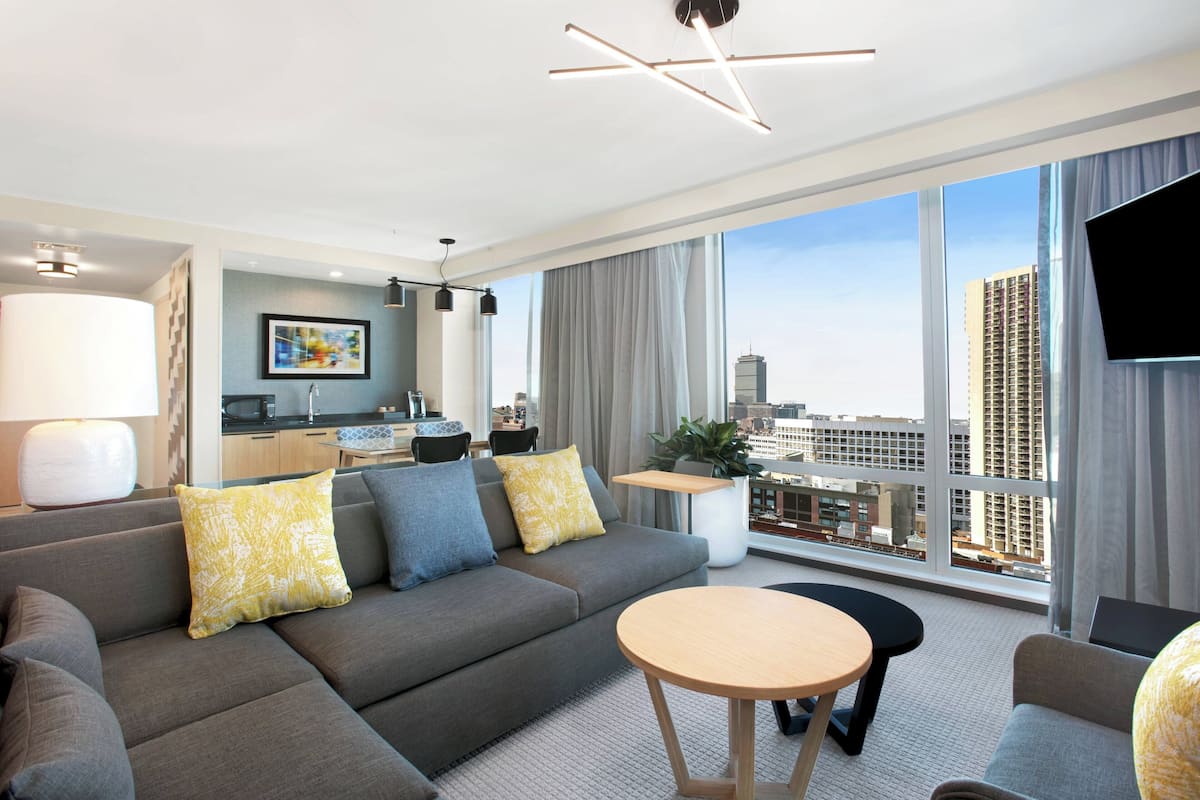 Een moderne hotelkamer met een grijze bank, een ronde houten tafel en uitzicht op de stad door grote ramen.