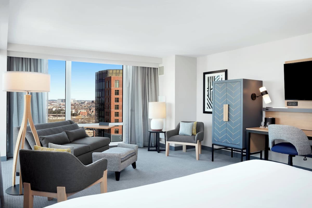 Een moderne hotelkamer met een groot bed, een grijze bank, een klein tafeltje, een bureau en een stadszicht door grote ramen.