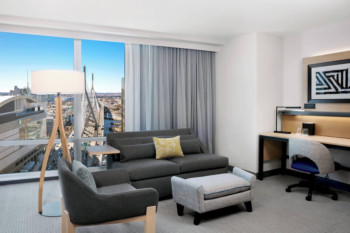 Een moderne hotelkamer met een grijze bank, een kleine tafel, een bureau met een computer en uitzicht op de stad door grote ramen.