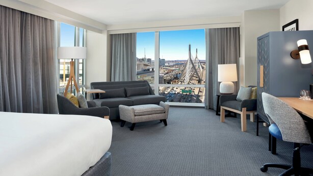 A hotel room with a large bed, a sofa, a desk, and a view of a cityscape with a bridge. - Courtyard by Marriott Boston Downtown/North Station (Boston)