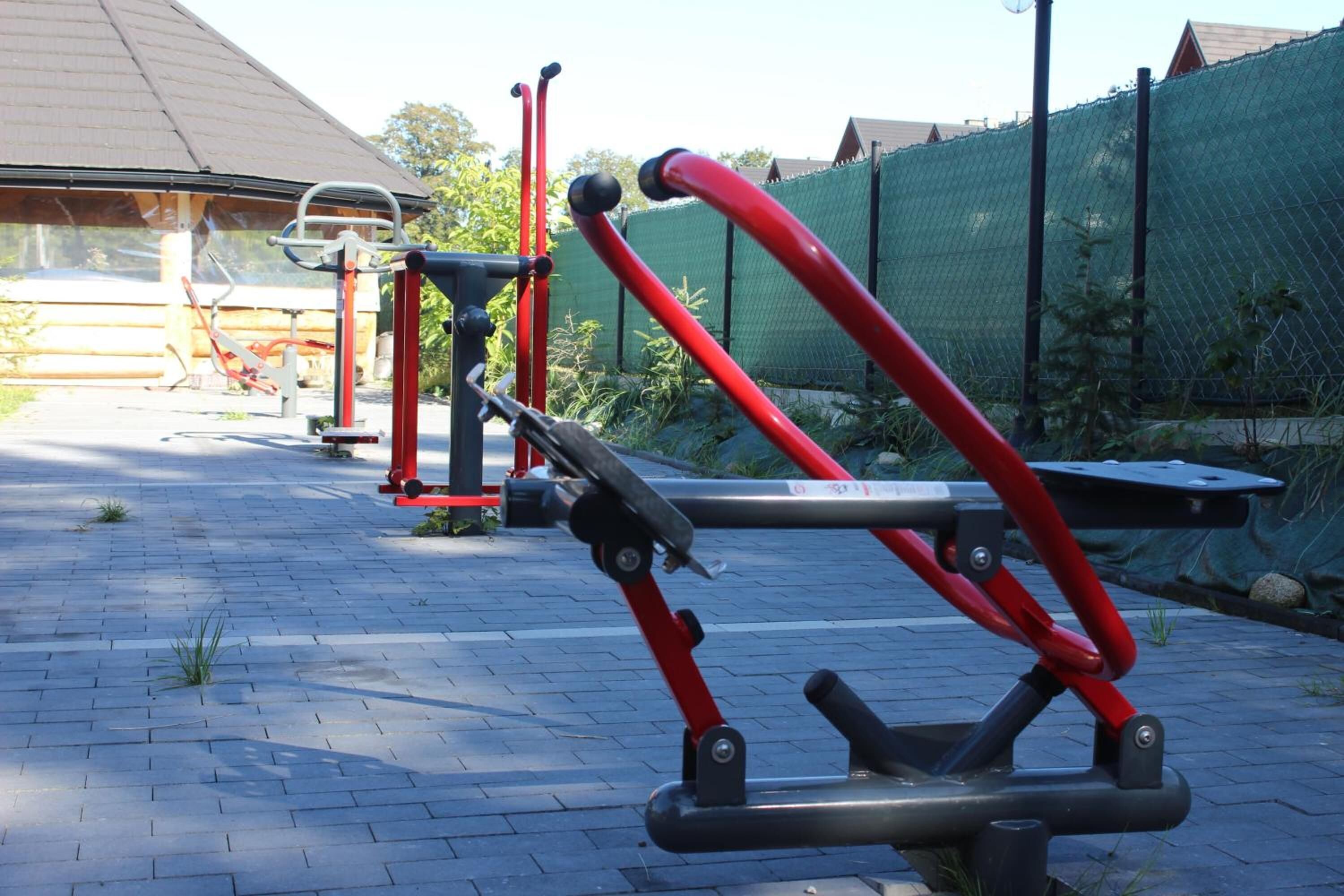 Outdoor exercise equipment in a paved area with a wooden building and a green fence in the background.