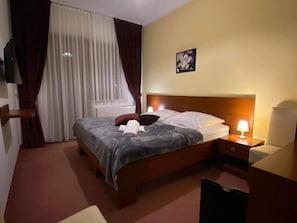 Habitación de hotel con cama, lámparas de noche, ventana con cortinas, televisor montado en la pared y un cuadro.