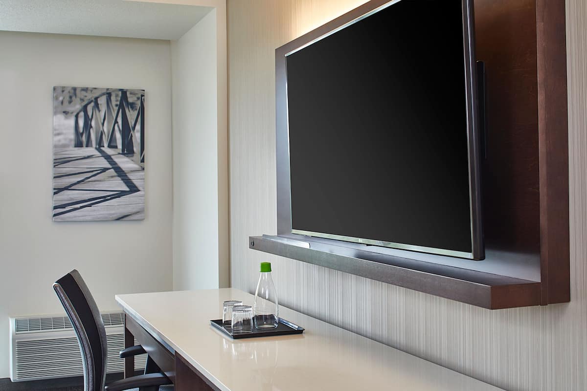 Habitación de hotel con televisor de pantalla plana montado en la pared, una mesa de comedor con sillas y una mesita con una botella y vasos.