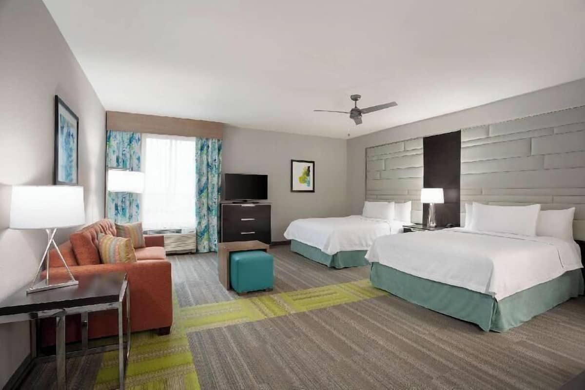 A hotel room with two beds, a sofa, a TV, a desk, and a ceiling fan.