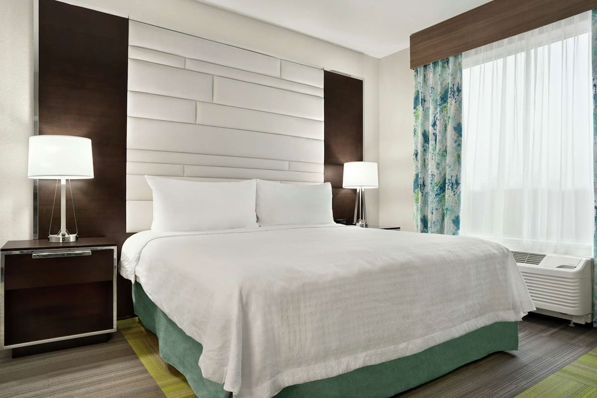 Habitación de hotel con una cama grande, dos mesitas de noche con lámparas, una ventana con cortinas y un aire acondicionado instalado en la pared.