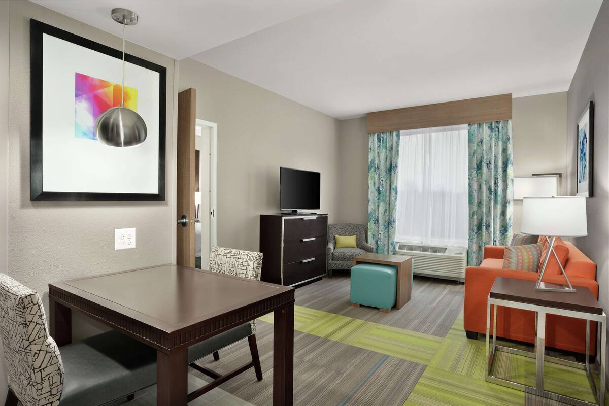 A hotel room with a dining table, sofa, TV, and a framed picture on the wall.