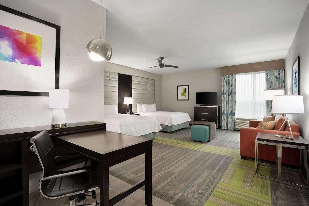 A hotel room with a desk, two beds, a sofa, a TV, and a ceiling fan.