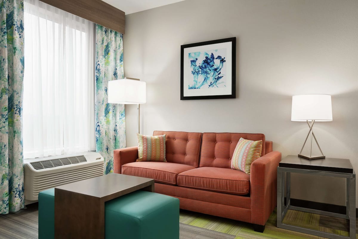 A hotel room with a sofa, a side table, a lamp, a framed artwork, and a window with curtains.
