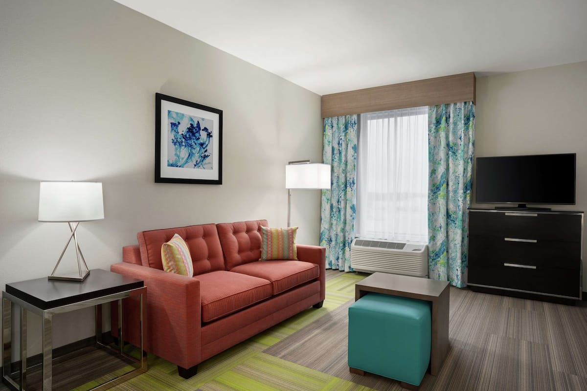 A hotel room with a red sofa, a teal ottoman, a TV, a desk with a lamp, and a framed picture on the wall.