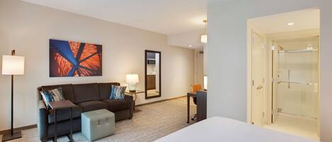 Habitación de hotel con cama, sofá, escritorio, espejo y cabina de ducha de vidrio.