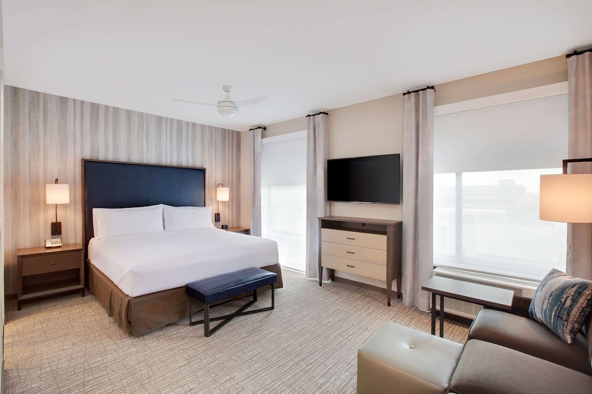 A hotel room with a bed, bedside tables, a desk with a flat-screen TV, a chair, and a window with curtains.