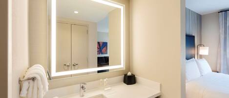 A hotel bathroom with a large mirror, white sink, and a bed with white linens.