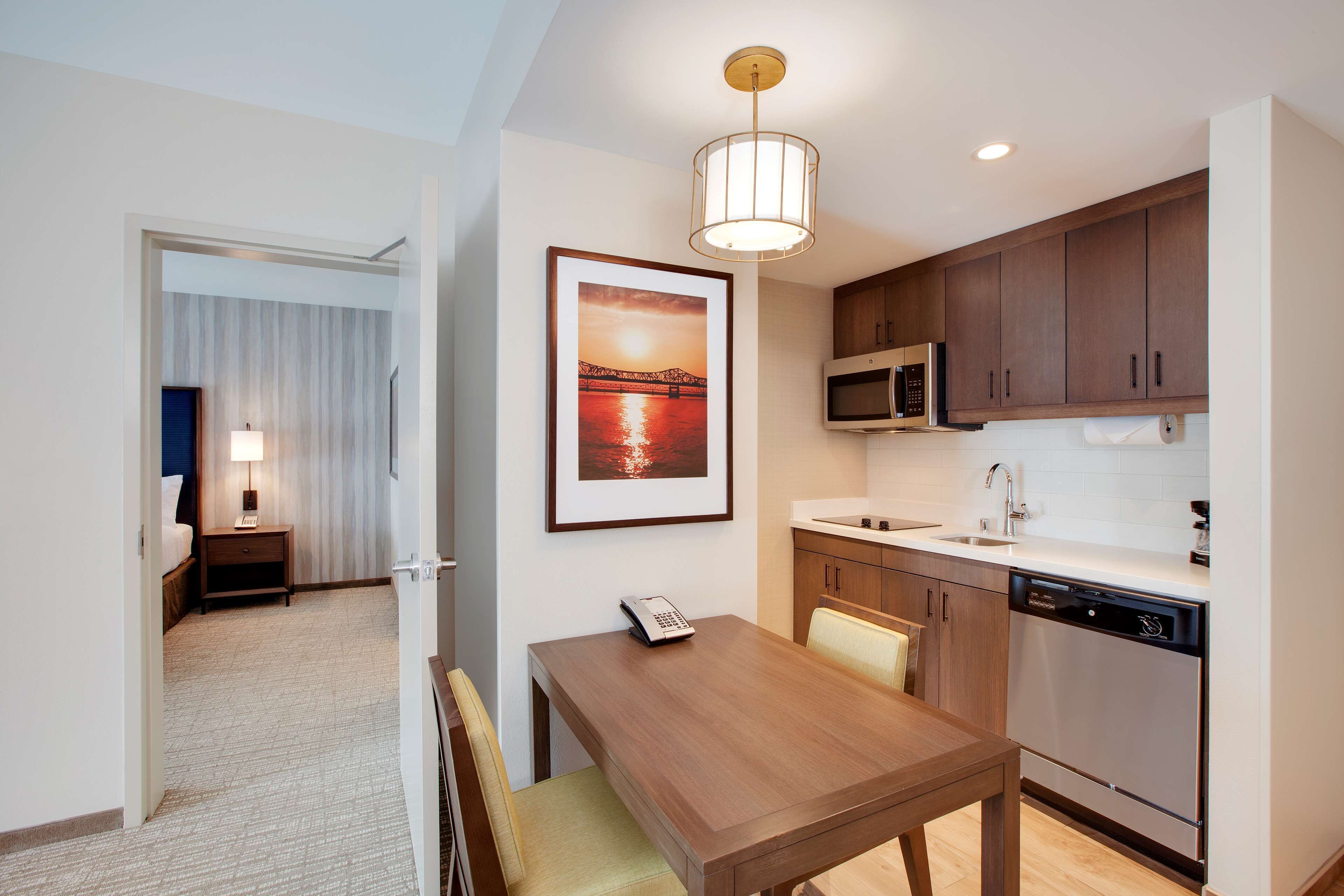 A hotel room with a wooden dining table, a microwave, a dishwasher, and a framed picture of a bridge.