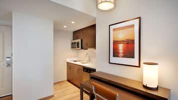 A modern kitchen with wooden cabinets, a microwave, and a sink. There is a framed picture of a sunset over a bridge on the wall, and a table with a chair and a floor lamp.