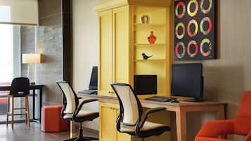 Un espace de bureau moderne avec un bureau jaune, deux chaises de bureau, un ordinateur et un écran. On y trouve une bibliothèque avec des objets décoratifs et un tableau accroché au mur.