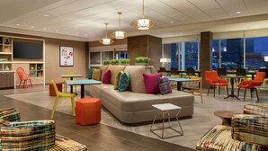 A modern lounge area with a sofa, colorful chairs, and tables, featuring a large window with a city view. - Home2 Suites by Hilton Frederick (Frederick)