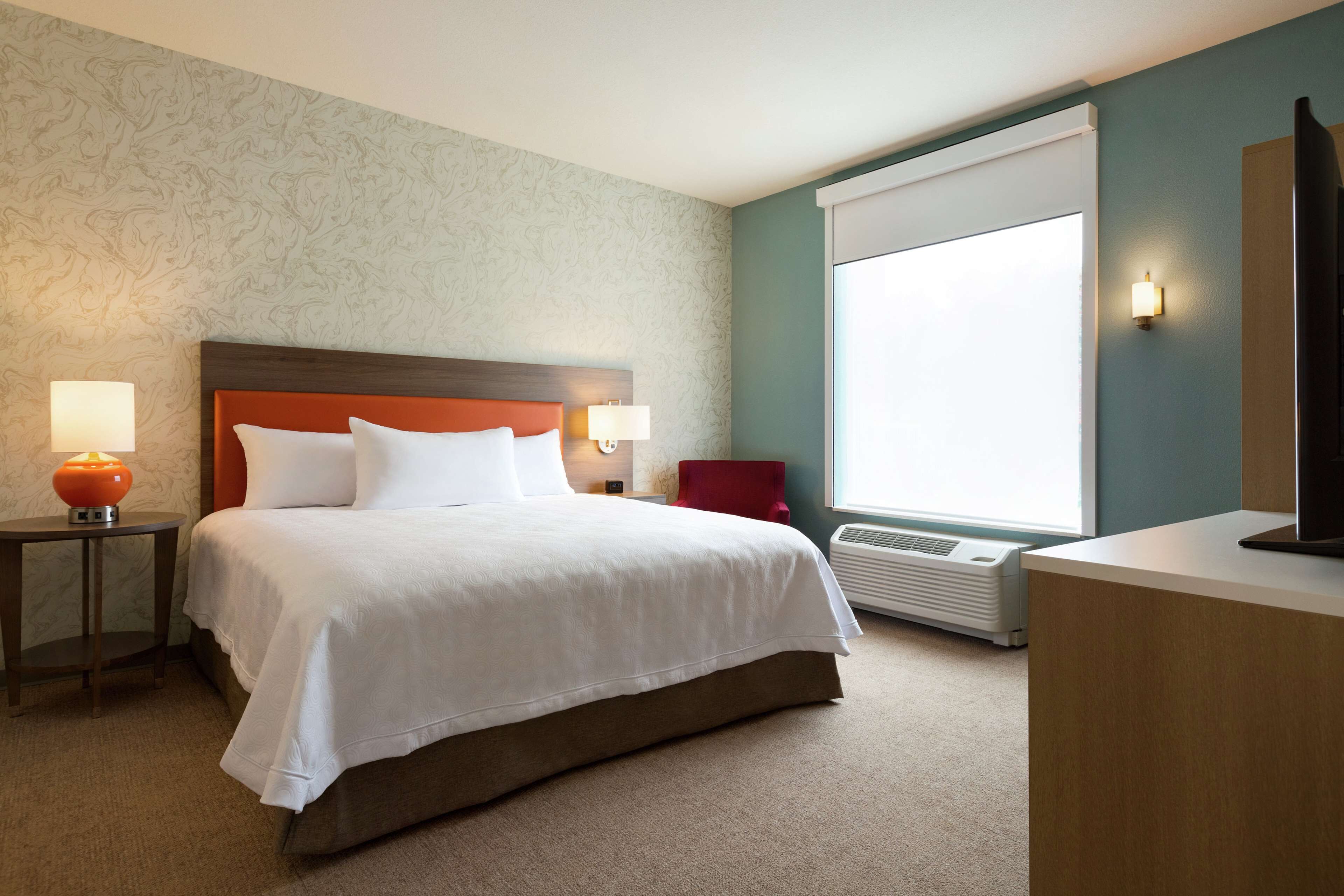 A hotel room with a large bed, two bedside lamps, a red chair, a desk, and a large window.