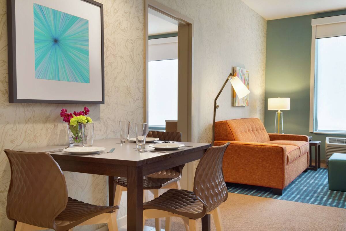A hotel room with a dining table set for two, a sofa, a lamp, and a window with a view.