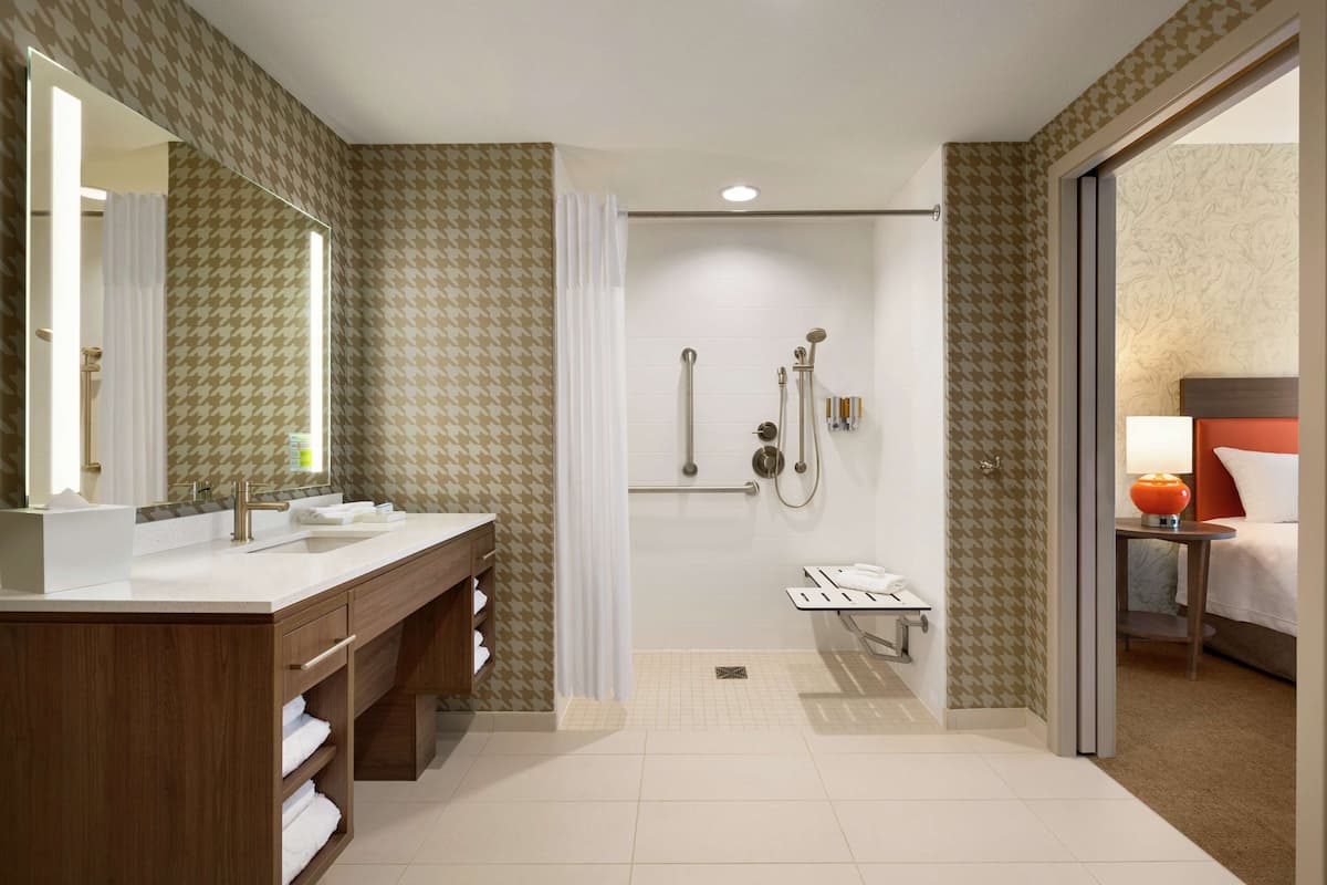A modern bathroom with a shower, vanity, and a bed visible through an adjacent room.