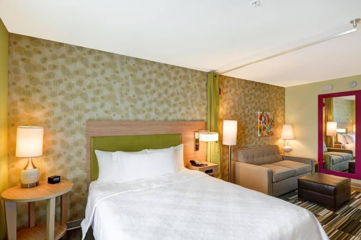 A hotel room with a bed, two bedside tables with lamps, a sofa, a mirror, and a carpeted floor.