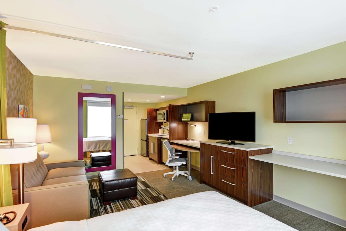 A hotel room with a bed, a sofa, a desk with a computer, a TV, and a bathroom visible through an open door.