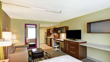 Quarto de hotel com cama, sofá, secretária com computador, televisão e banheiro visível por uma porta aberta.